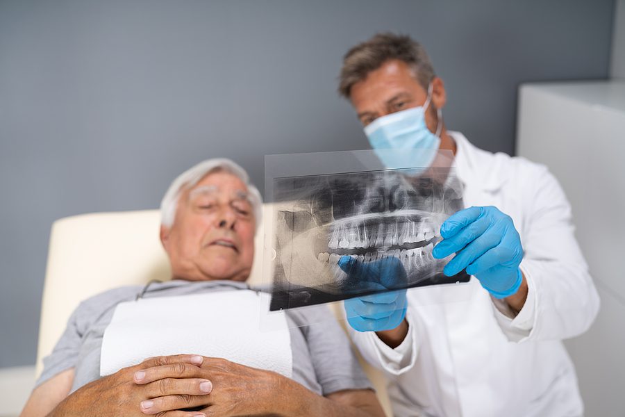 Dentist wearing a white coat and holding up dental X-rays to an older male patient. 
